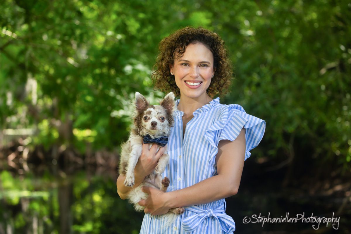 Smiling woman in blue striped dress holding a small Chihuahua mix dog outdoors among green trees.