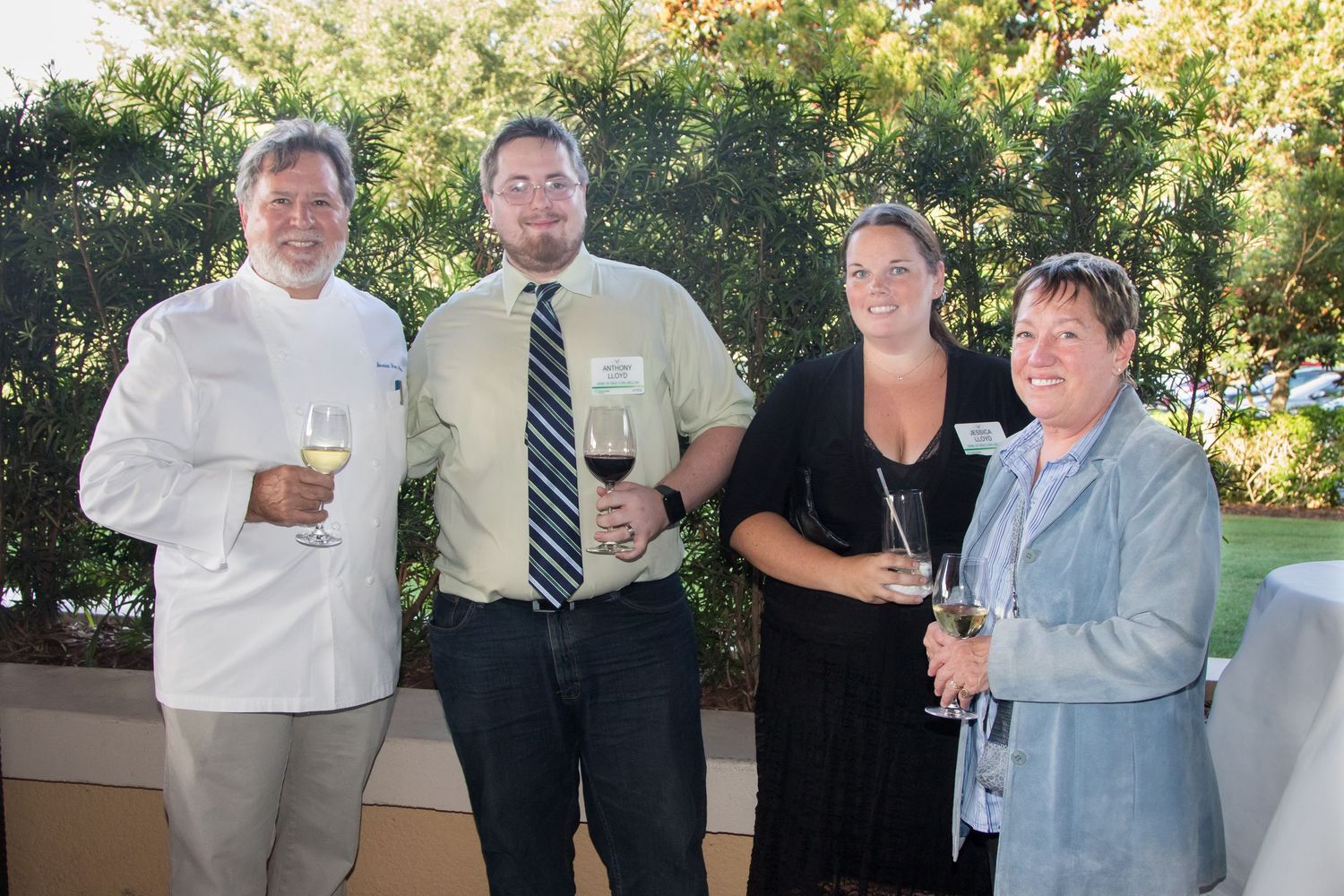 Four guests smile holding wine glasses at outdoor evening reception event.