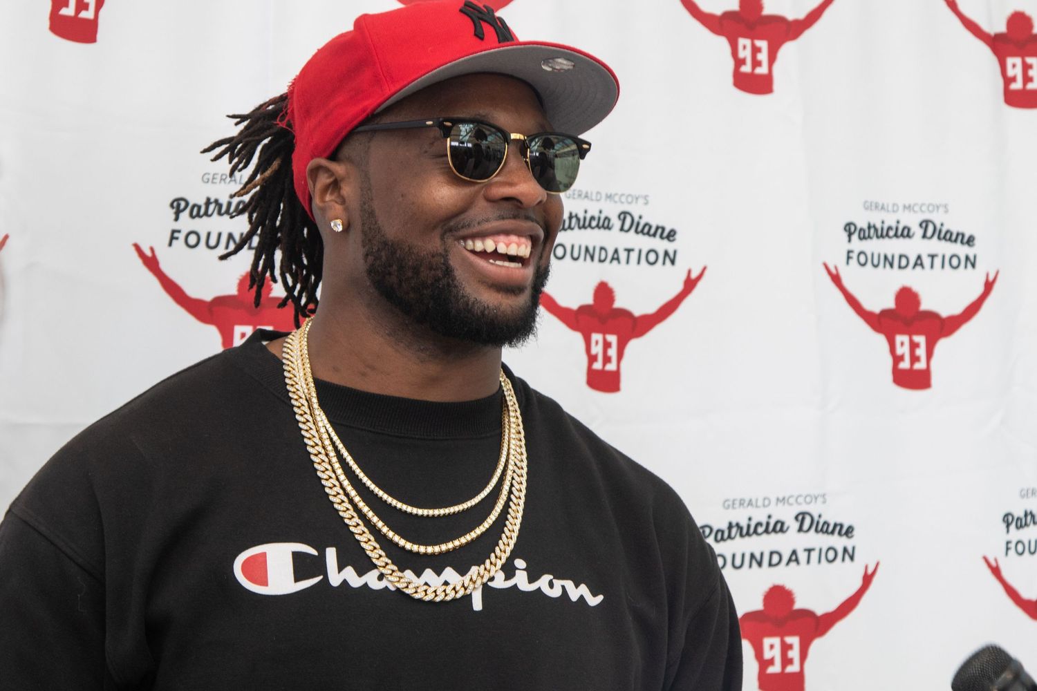 Smiling man in red Champion shirt and gold chains at Patricia Diane Foundation event backdrop.
