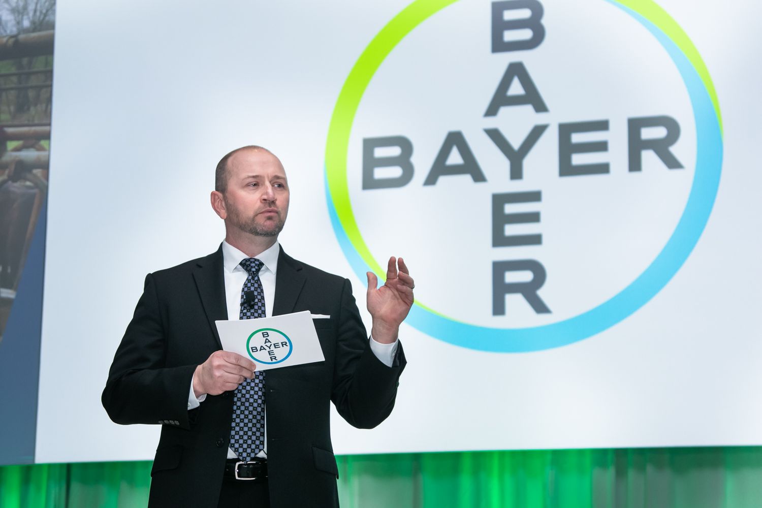 Professional speaker presents at Bayer corporate event holding papers before branded logo backdrop.
