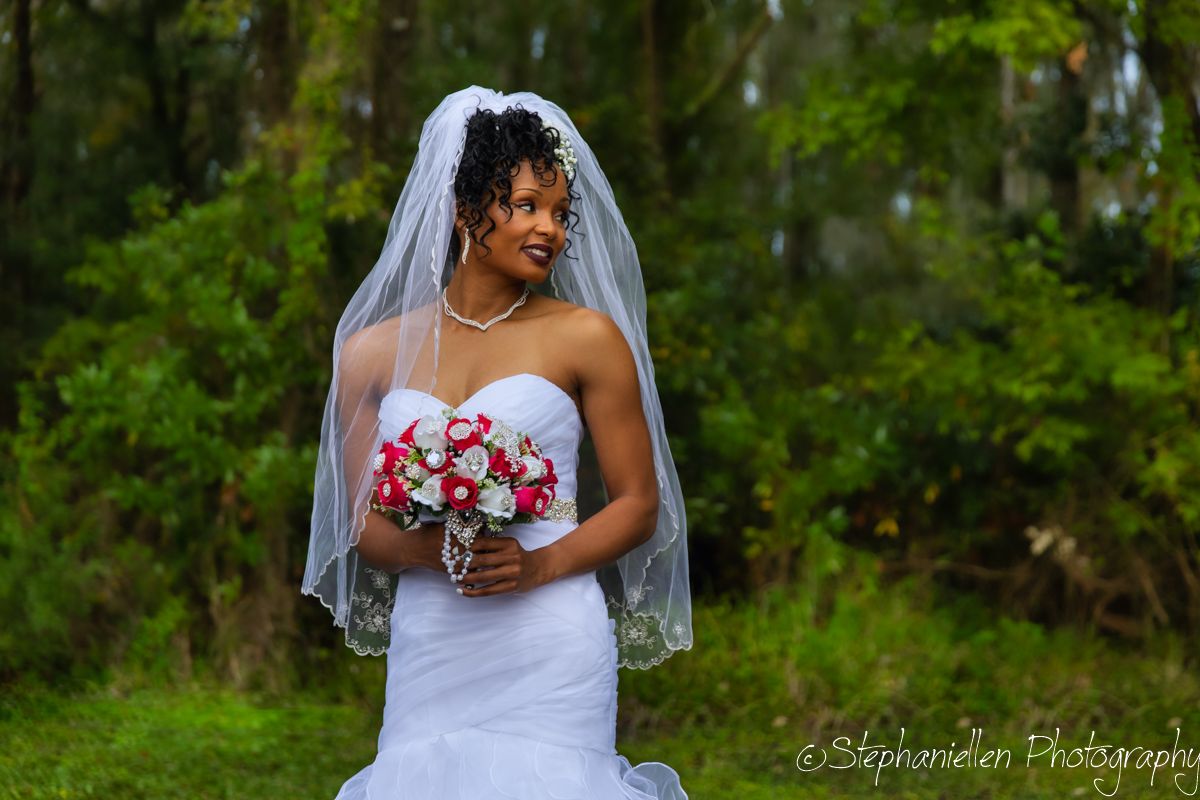 Beautiful Black bride in a white mermaid gown holding a red and white bouquet wearing a veil outdoors.