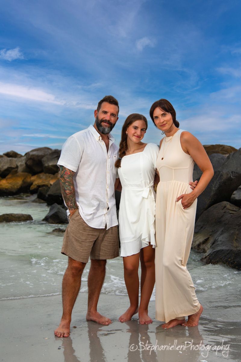 Family of three poses on a beach with rocky backdrop and blue sky, wearing white and cream outfits for a professional photo.