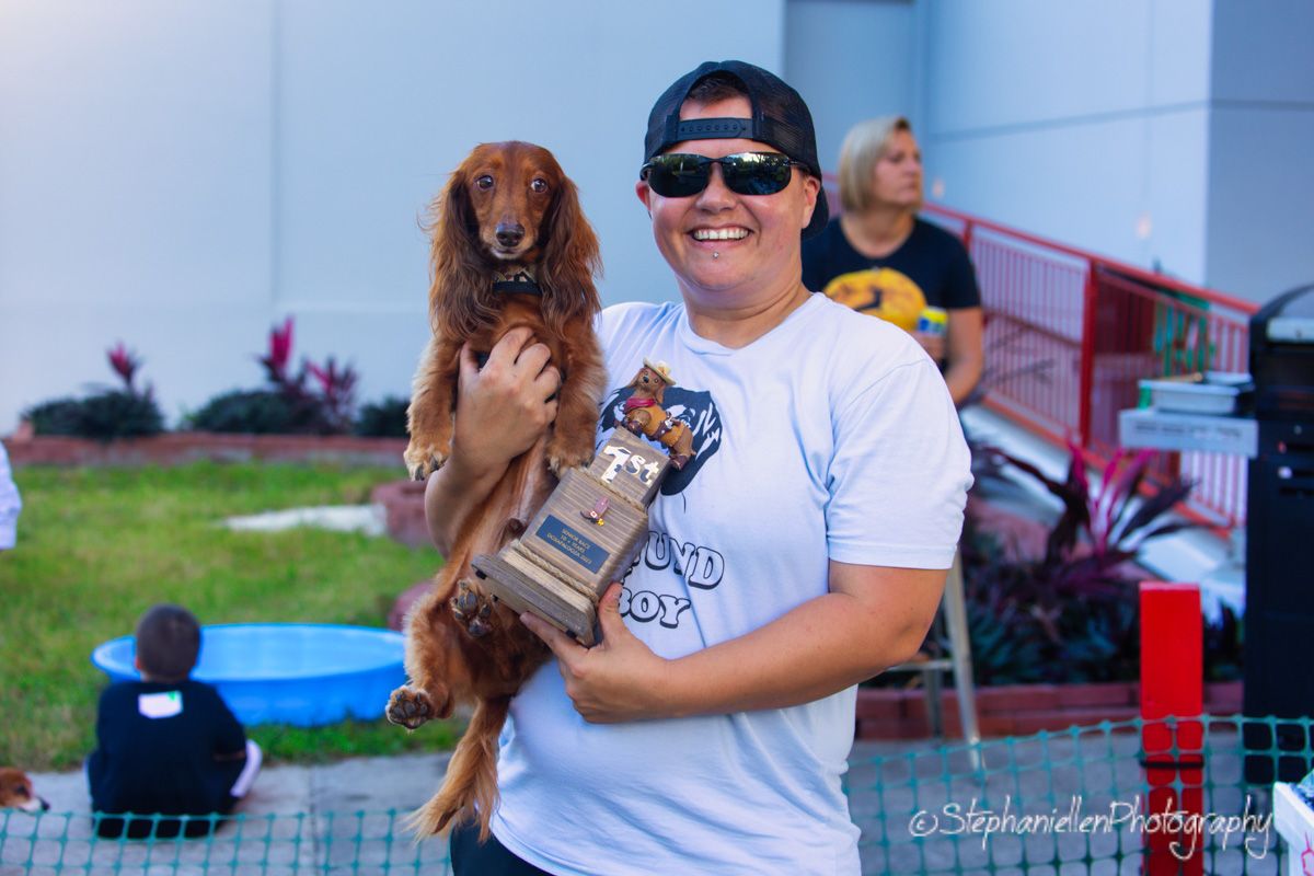Person holding a red dachshund and trophy at an outdoor dog show event, smiling proudly.