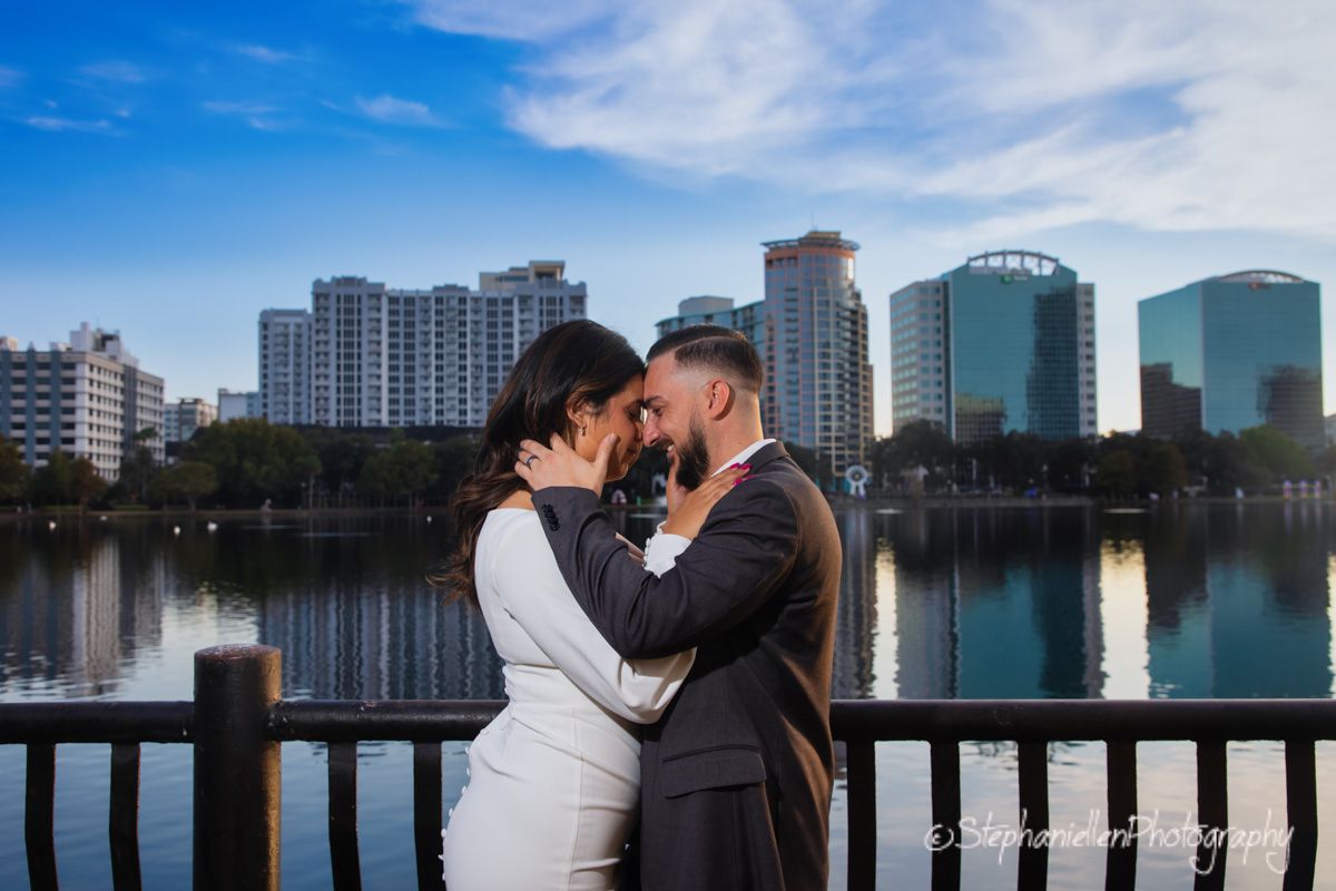 Couple sharing a kiss near a lake with city skyline and blue sky in background.