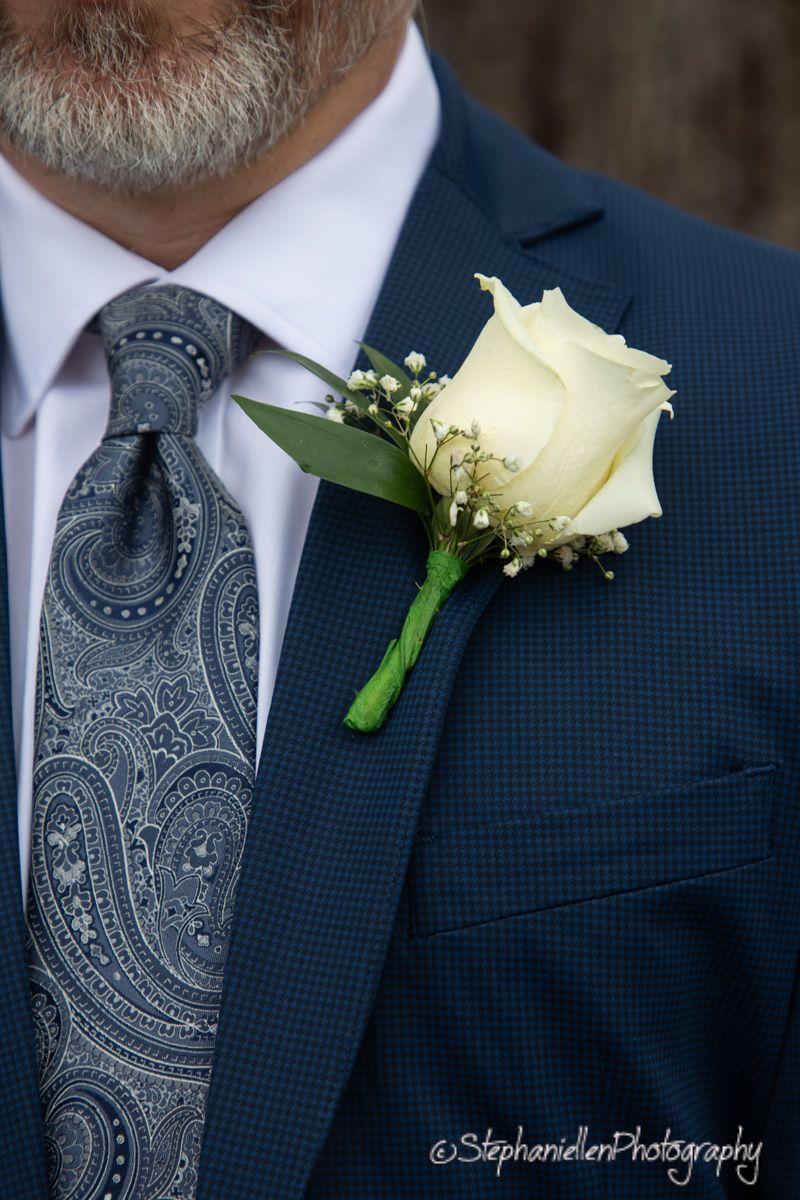 Groom wears white rose boutonniere on navy suit with gray paisley tie at wedding.