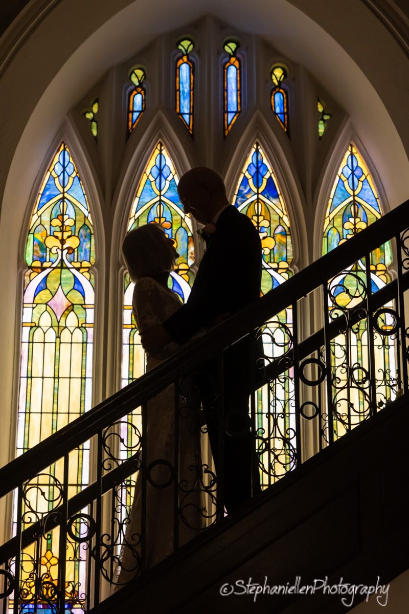 Bride and groom silhouetted against stunning stained glass windows on church staircase.