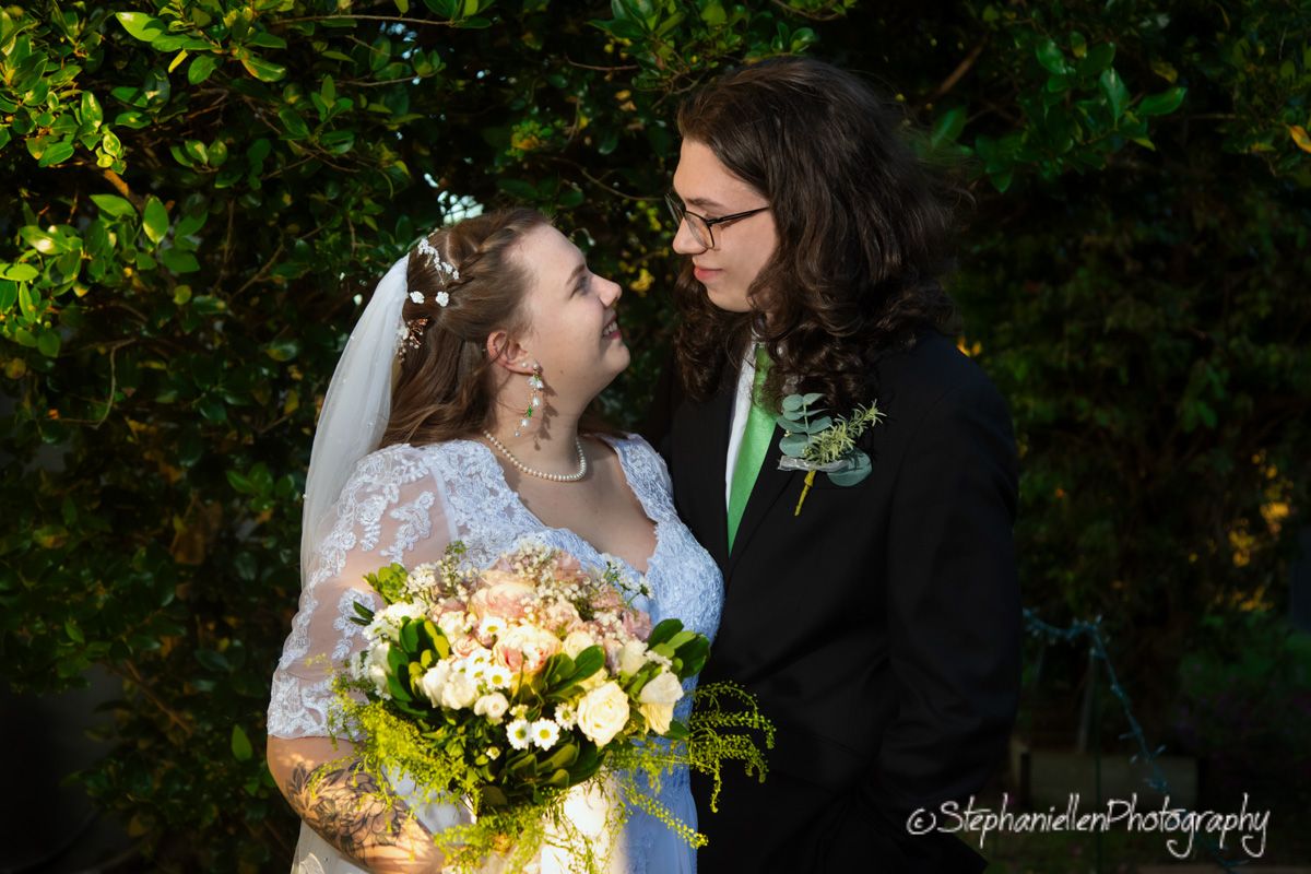 Two brides share a romantic moment outdoors, one in a lace gown with veil holding a bouquet, the other in a black suit.
