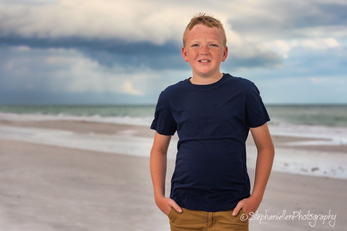 A smiling boy in a navy t-shirt and khaki pants poses on a beach with stormy skies and ocean waves behind him.