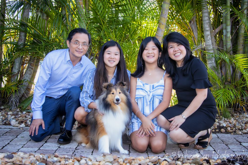 Family of four posing outdoors with a Shetland Sheepdog among tropical palms on a stone path.