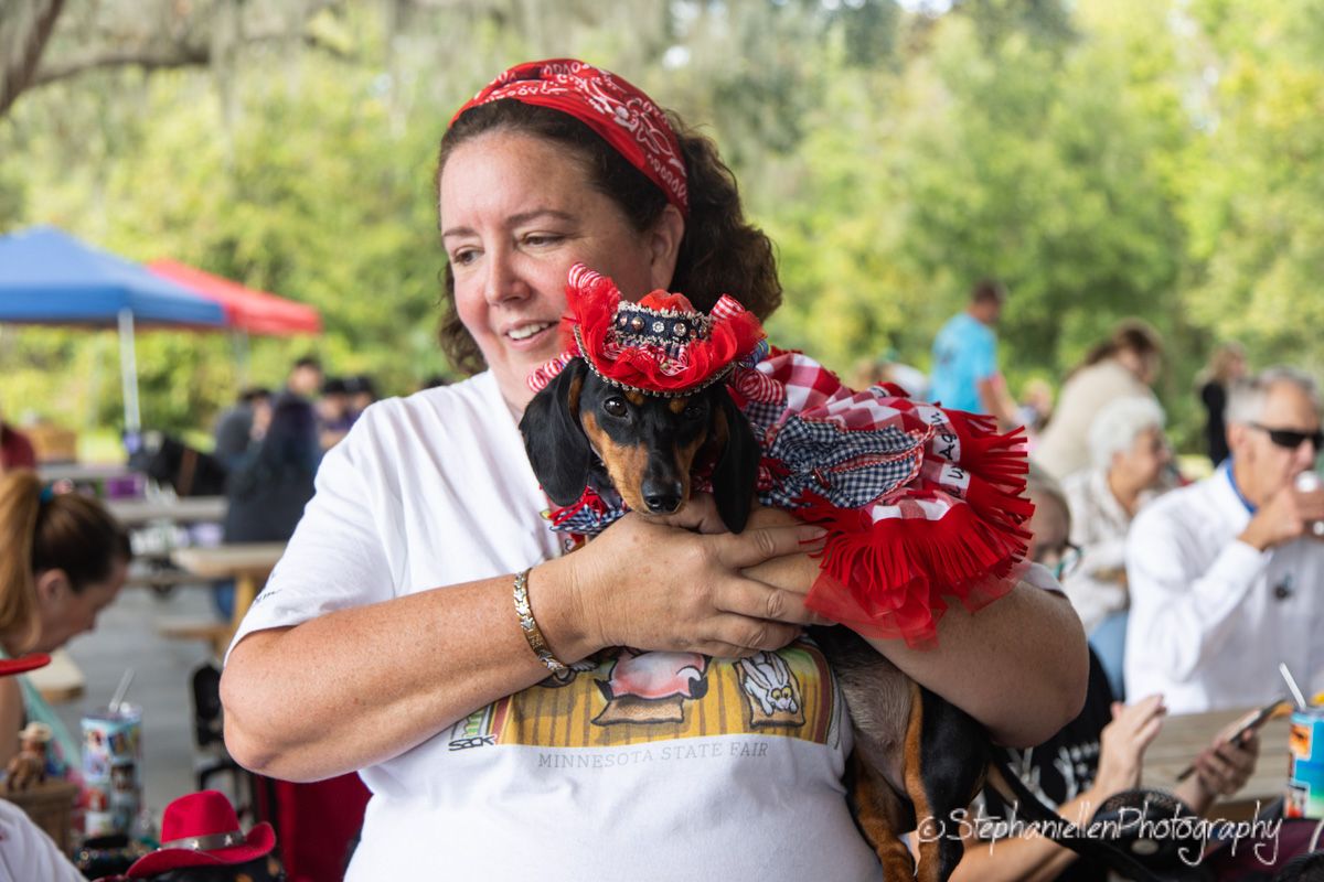 A smiling woman holds a dachshund dressed in a red bandana costume at an outdoor event.