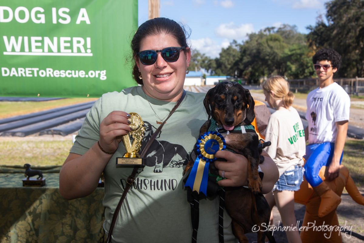 Woman holding a dachshund dog with a blue ribbon at an outdoor DAREToRescue.org wiener dog event.