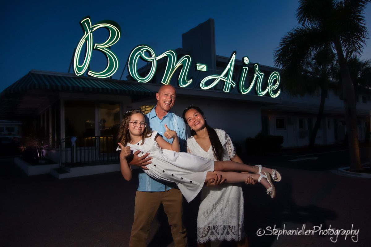 Family of three posing in front of glowing Bon-Aire neon sign at dusk.