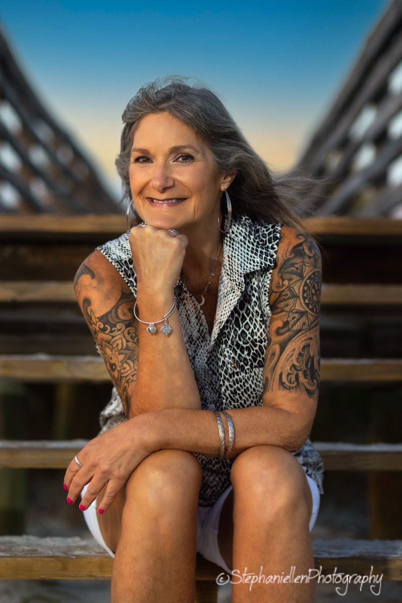 Confident mature woman with tattoos and gray hair smiling warmly while seated on a wooden bridge.