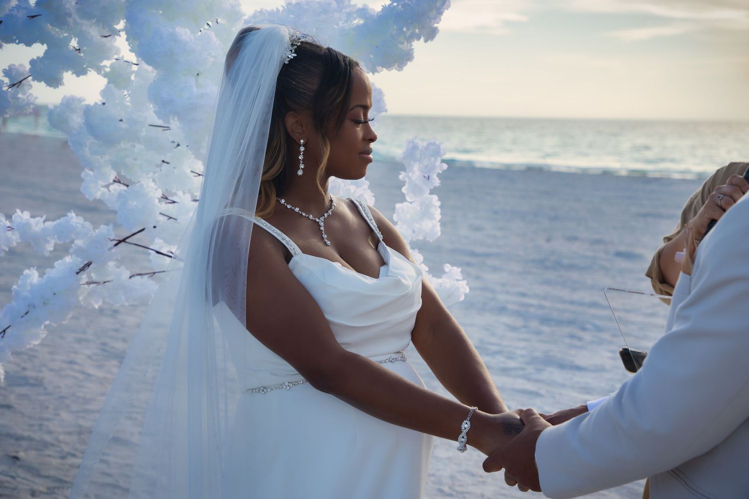 Bride in white gown and veil holding hands during beach wedding ceremony.