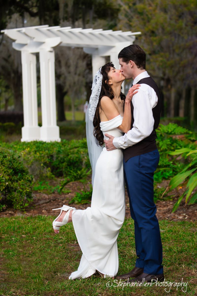 Bride and groom share romantic kiss near white pergola in garden.