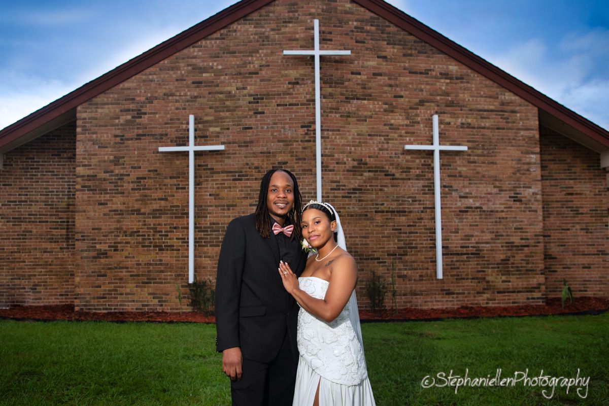 Bride and groom pose together in front of a brick church with white crosses.