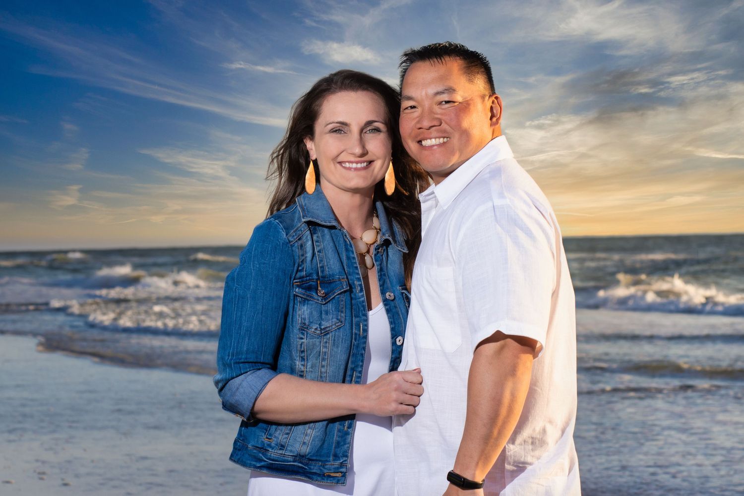 Couple smiling together on a beach at sunset, woman in denim jacket and man in white shirt.