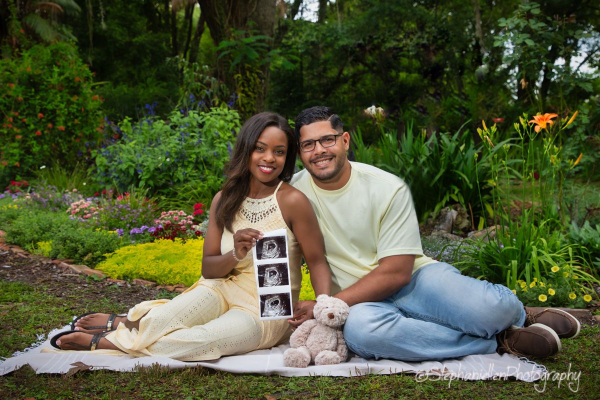 Happy couple sitting on a blanket in a garden, proudly displaying ultrasound photos of their baby.