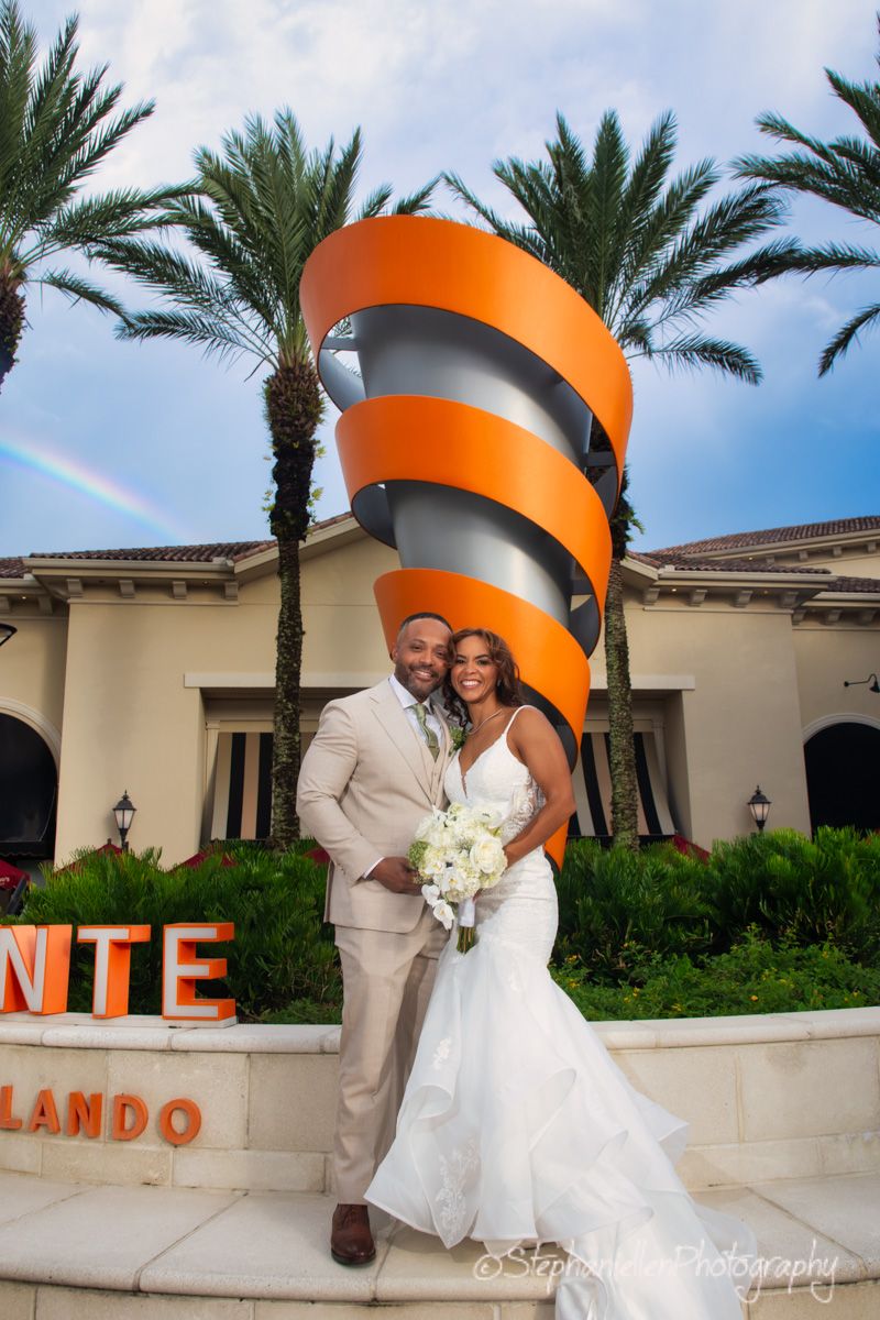 Wedding couple poses before a large orange spiral sculpture with a rainbow above.