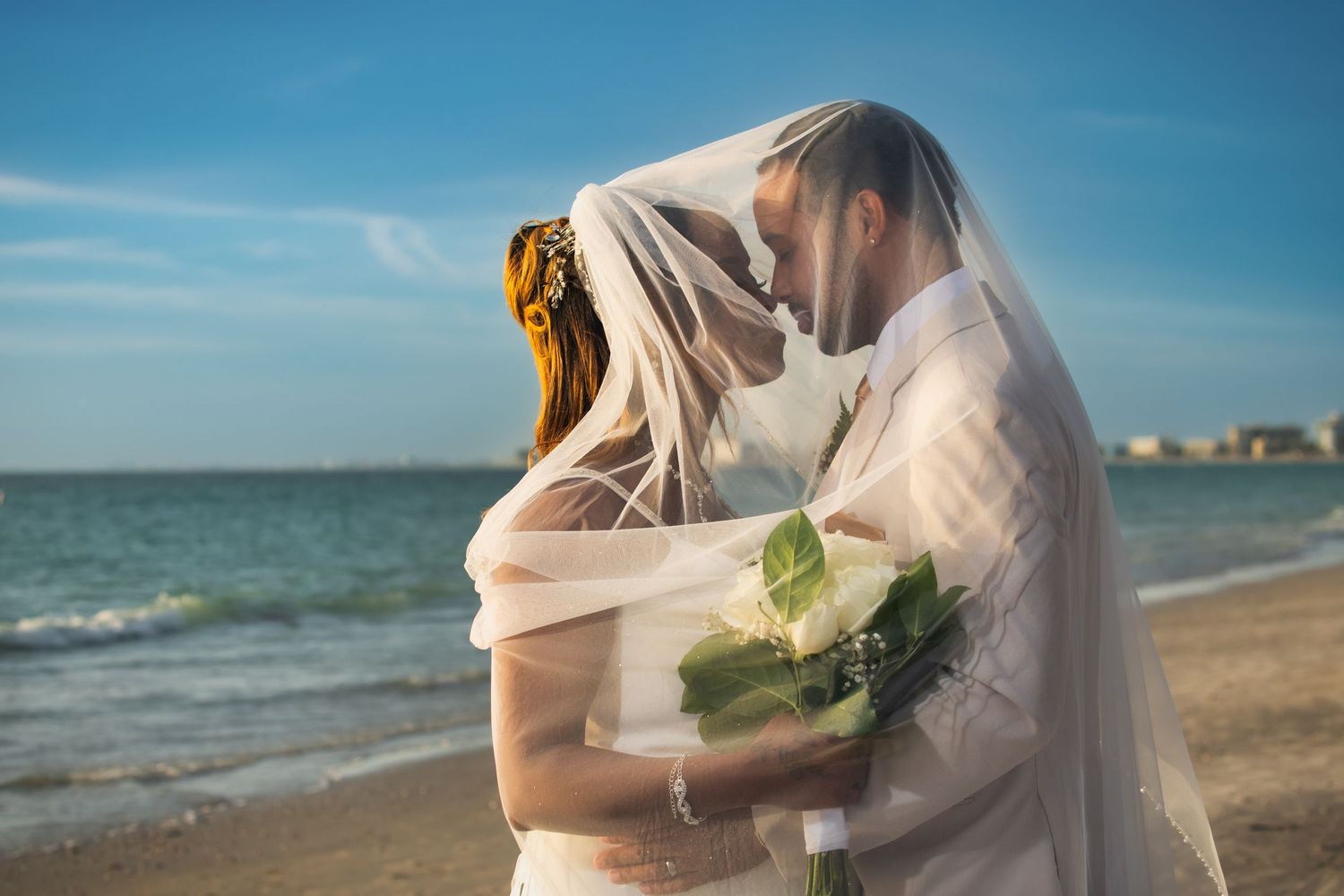 Bride and groom sharing a kiss under bridal veil on sandy beach.