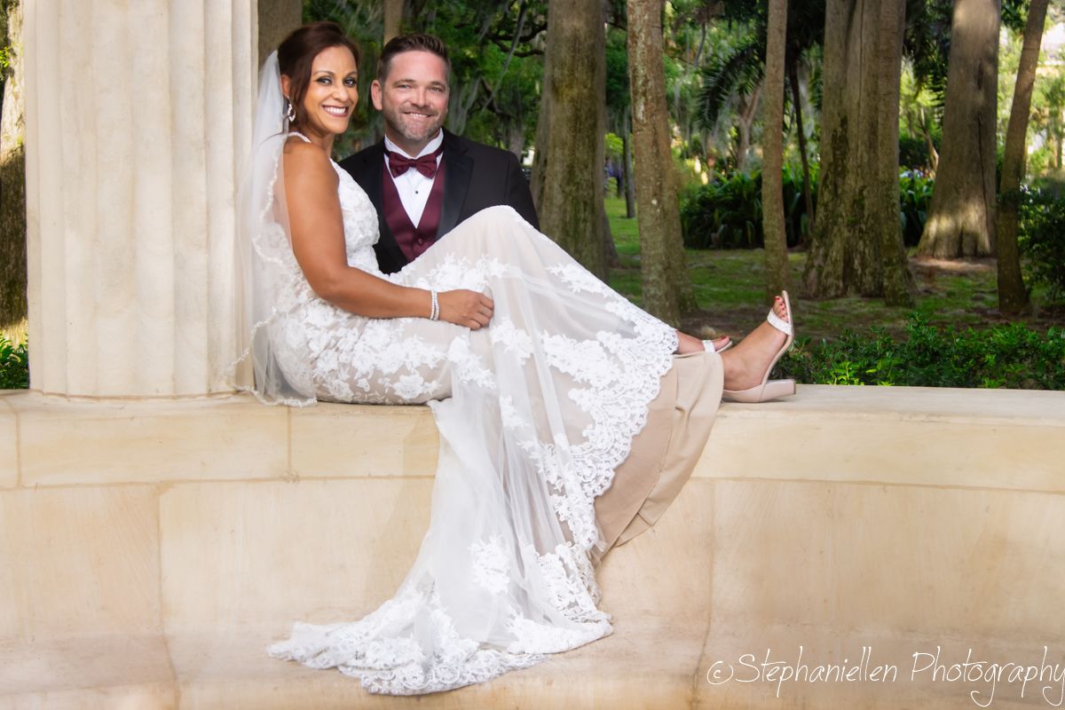 Groom in burgundy bow tie holds bride in lace wedding gown while seated on stone steps in park setting.