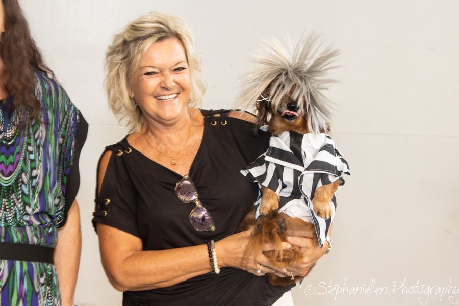 Woman holding a small dog dressed in a zebra costume with a wild gray wig at a pet event.