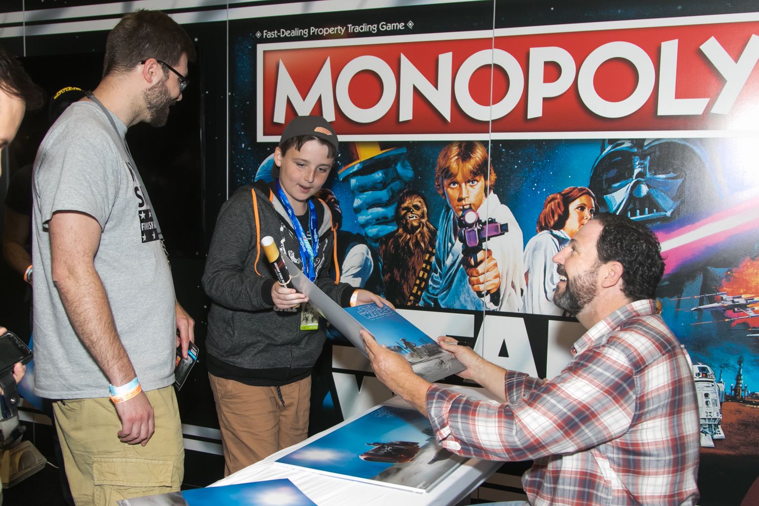 Man signs Star Wars Monopoly memorabilia for fans at convention merchandise booth.
