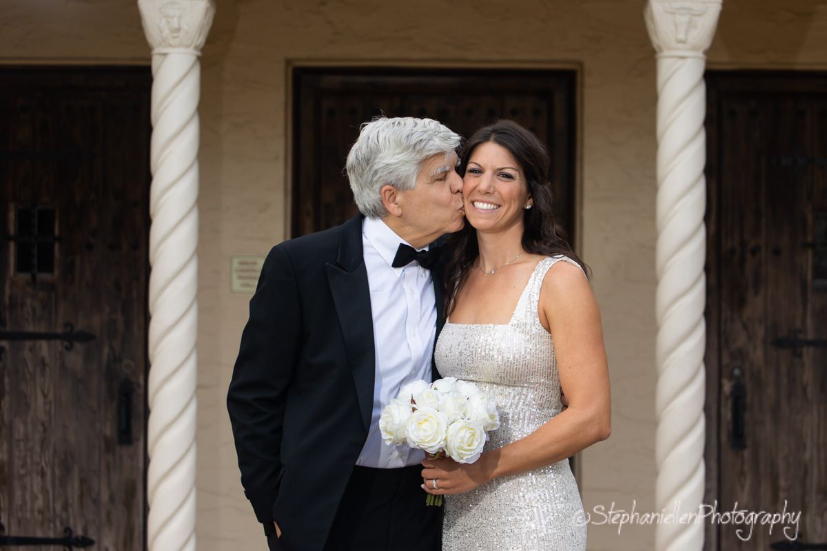A groom in a tuxedo kisses his brides cheek as she smiles holding white roses before a rustic wooden door.