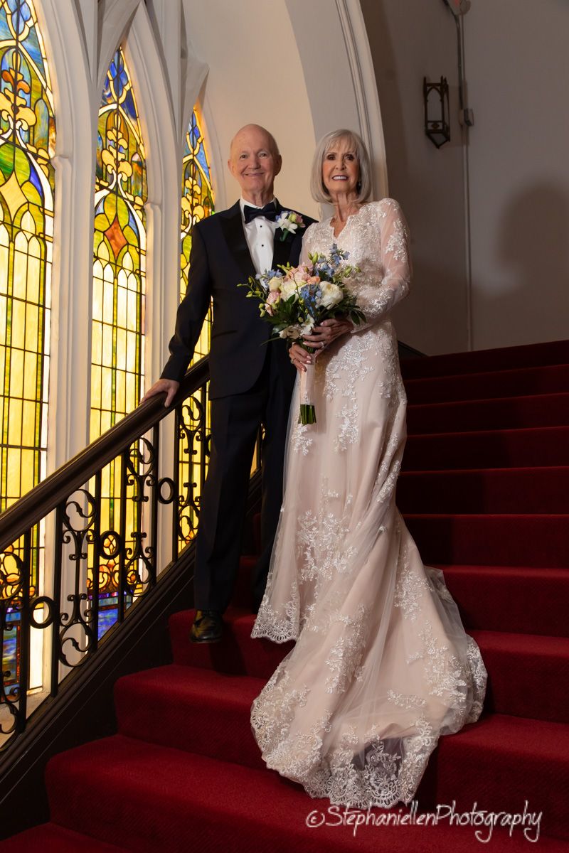 Bride in lace gown and groom in tuxedo descend red-carpeted church stairs with bouquet.