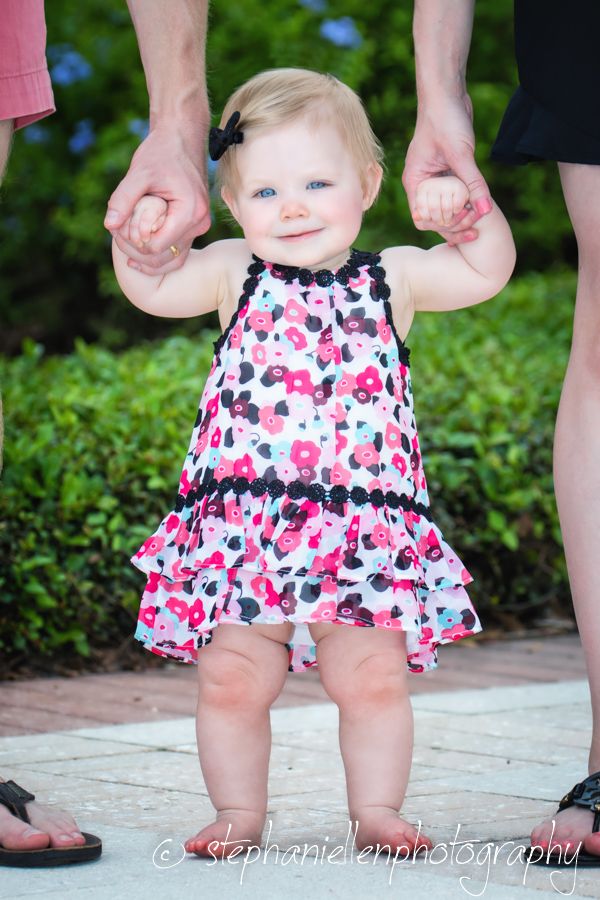 A smiling baby girl in a pink floral dress stands outdoors, held up by two adults hands on a stone path.