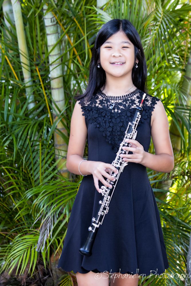 Young girl in black dress smiling while holding an oboe outdoors among tropical palm trees.