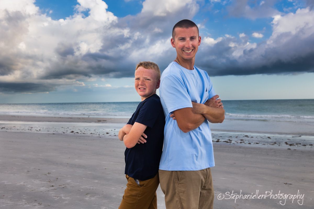 A man and young boy stand back-to-back with arms crossed on a beach, smiling under a dramatic cloudy sky.