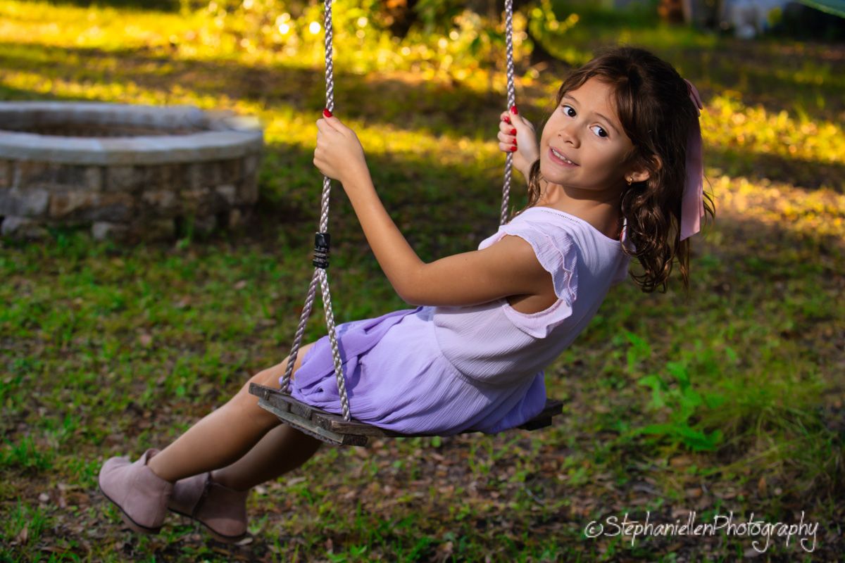 Young girl smiling on a rope swing outdoors in golden sunlight, wearing a purple skirt and white top.