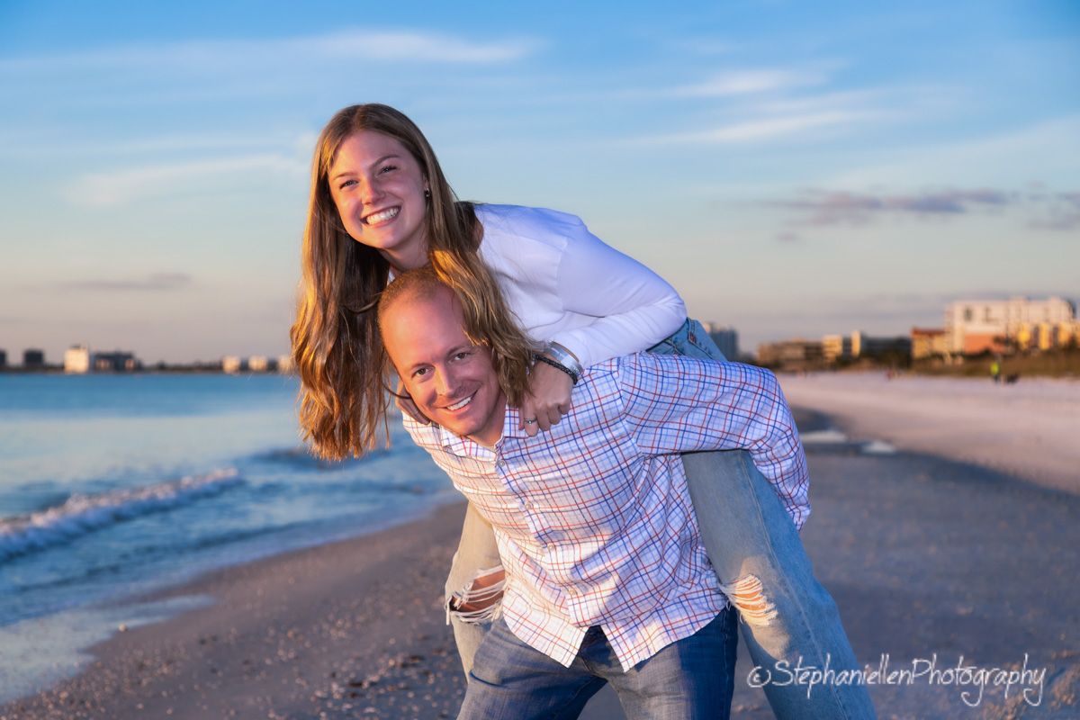 A young couple laughs joyfully on a sunny beach, the man giving the woman a piggyback ride near the ocean.