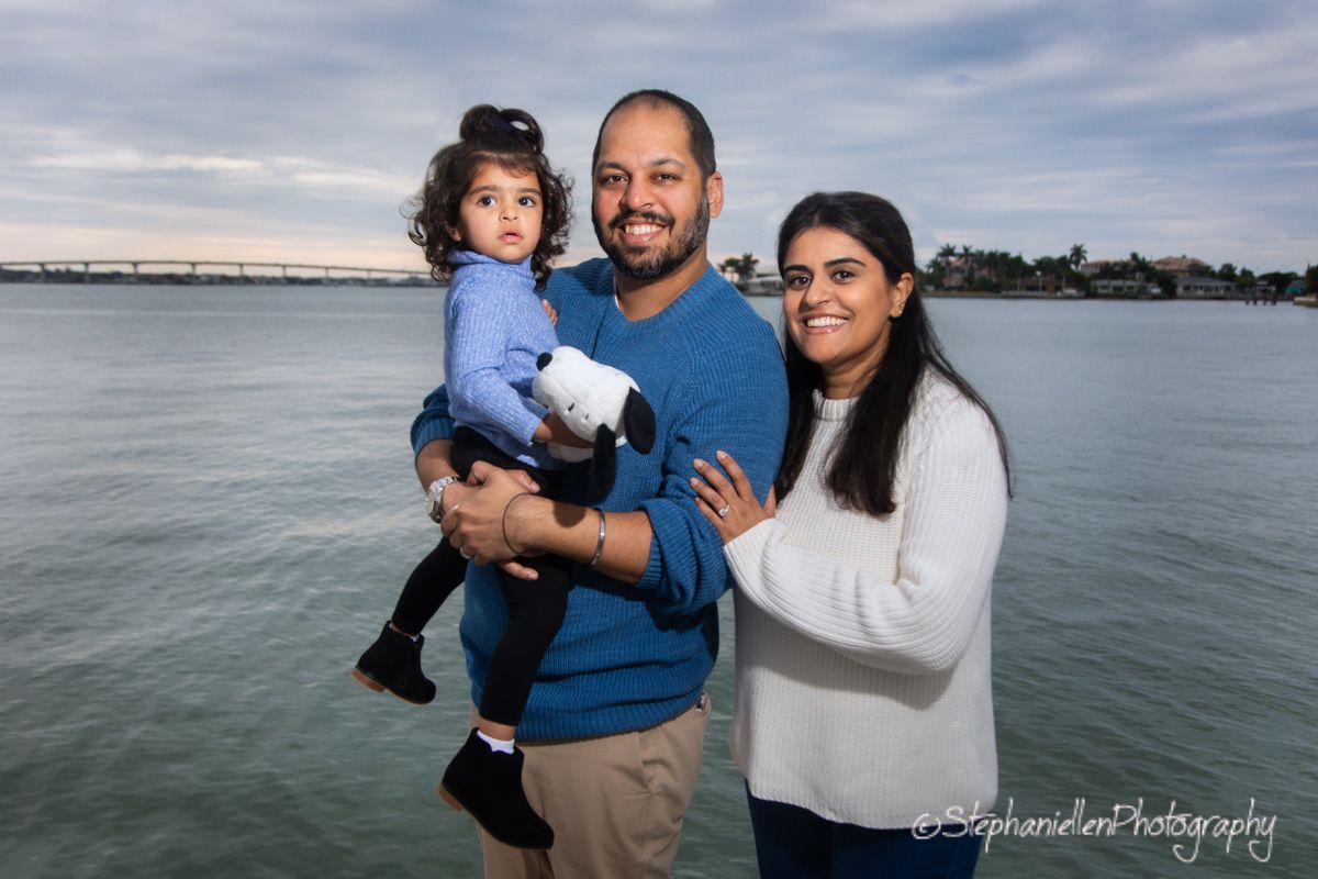 A happy family of three poses by the waterfront, with a man holding a toddler and a woman smiling beside them.