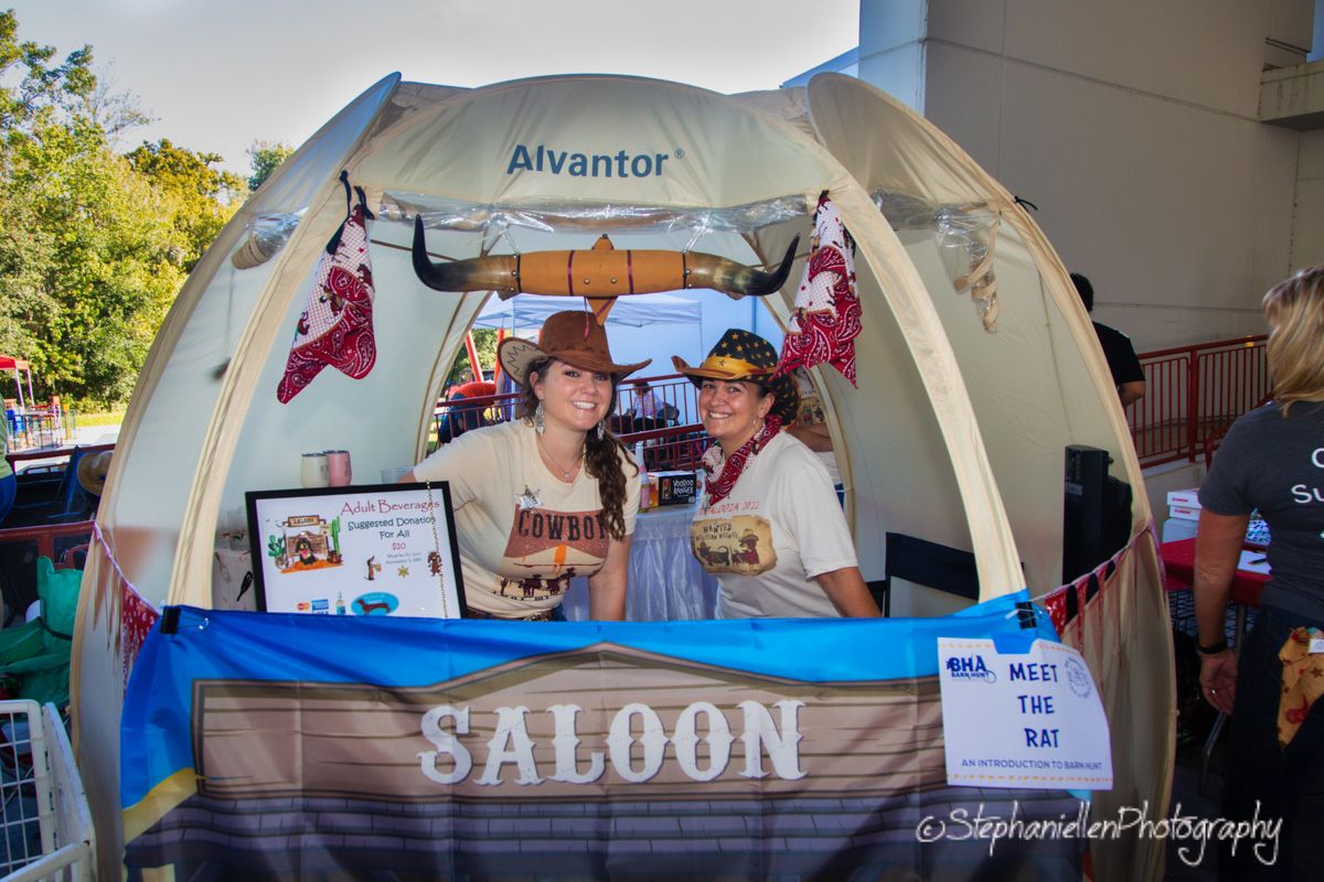Two women in cowboy hats smile inside an Alvantor tent decorated as a Western saloon booth at an outdoor event.