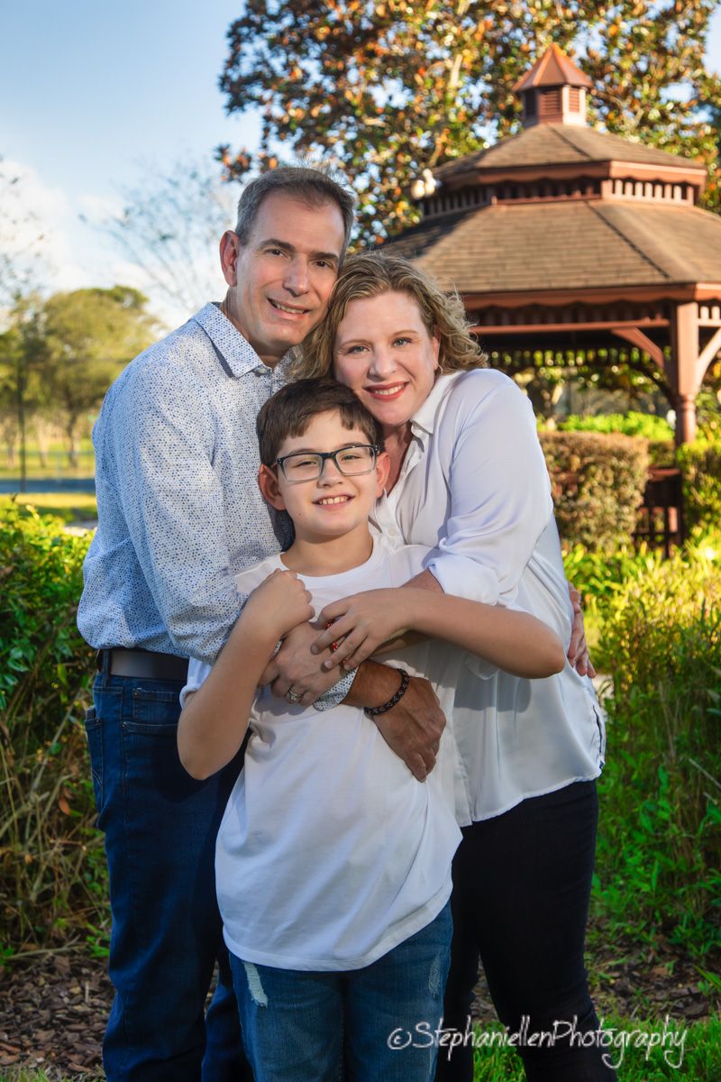 Happy family of three posing outdoors near a gazebo, parents embracing their young son in casual attire.