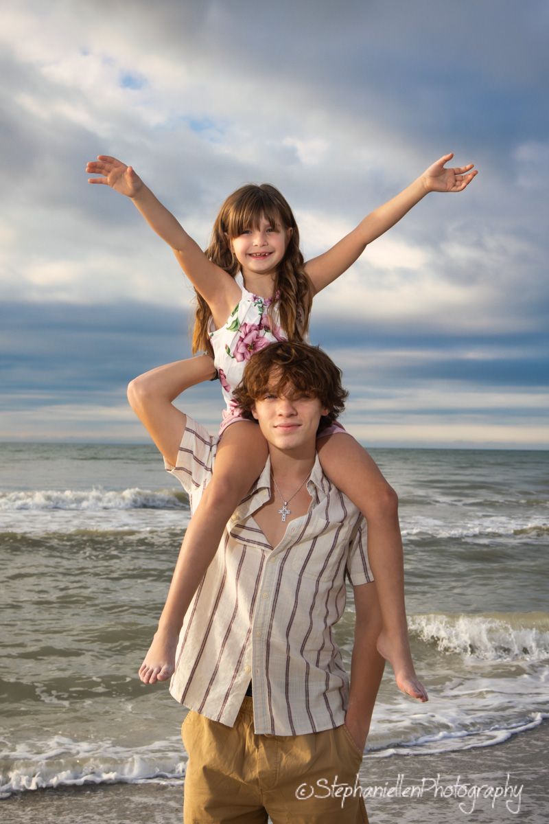 A teenage boy carries a young girl on his shoulders at the beach, as she spreads her arms wide with joy.