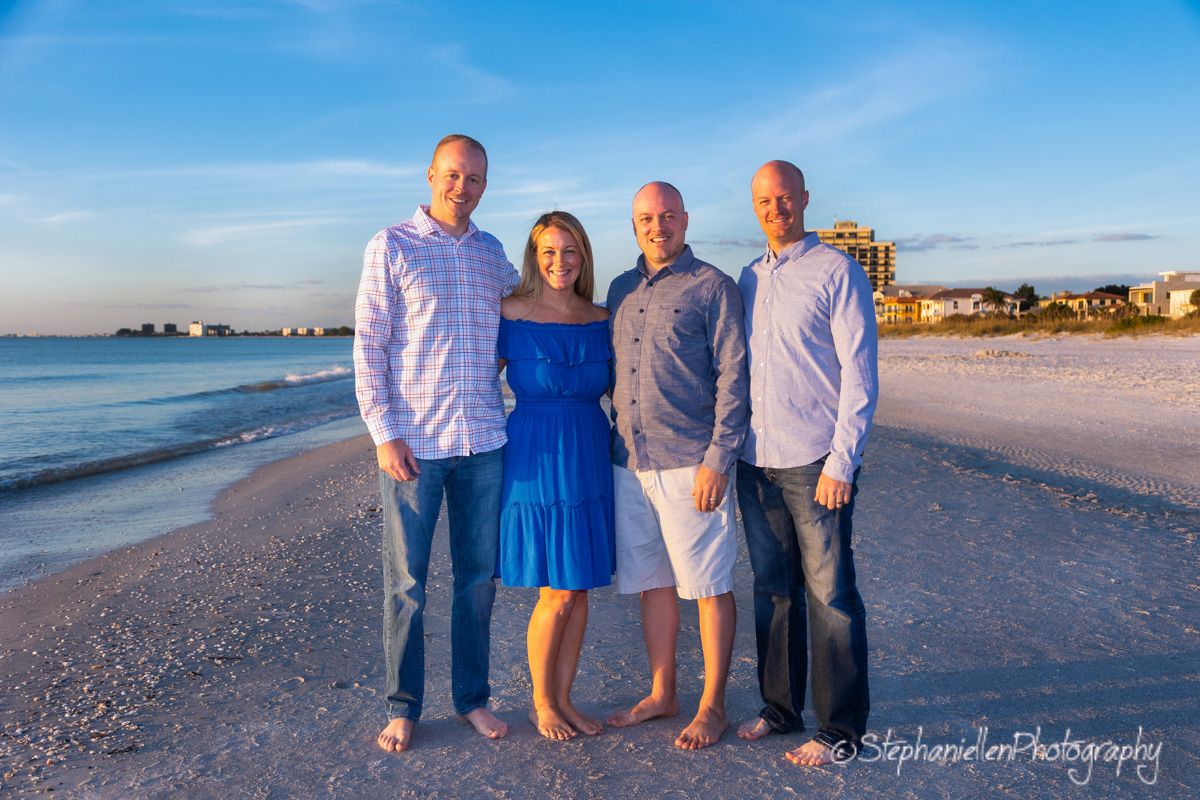 Family of four poses together on a sunny Florida beach with blue sky and coastal buildings in the background.