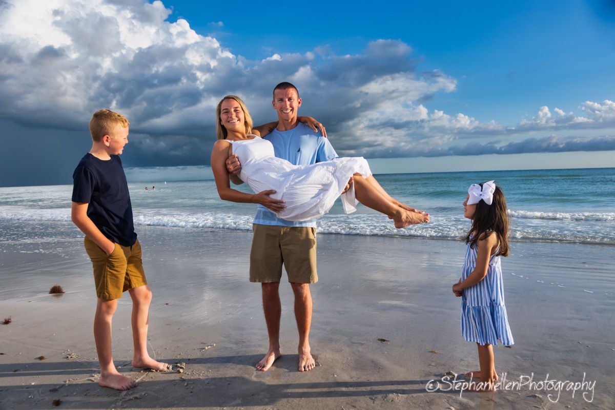 Family beach photo with man carrying woman while two children stand nearby, ocean waves and dramatic cloudy sky behind them.