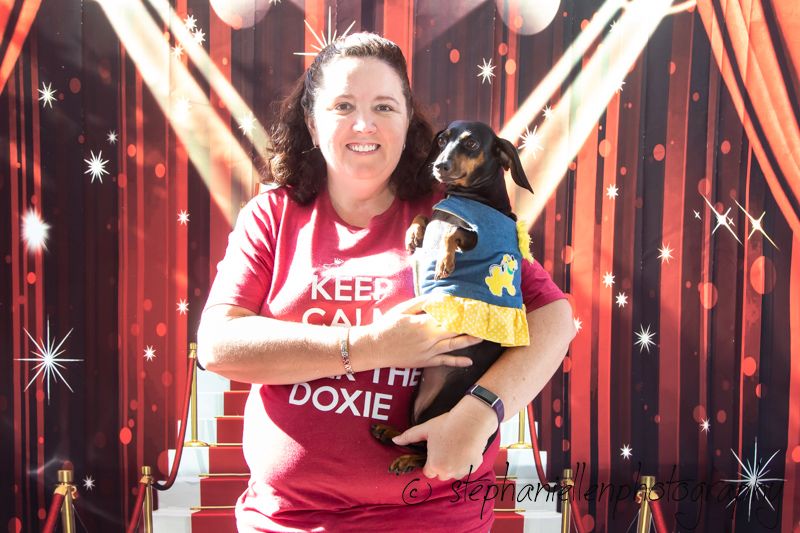 Woman in red Keep Calm dachshund shirt holds small dog in yellow outfit before red carpet backdrop.