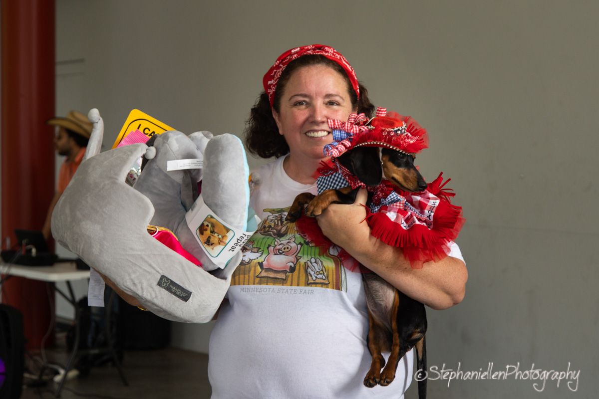 Woman smiling holding a costumed dachshund in red outfit and a stuffed elephant toy at an outdoor event.