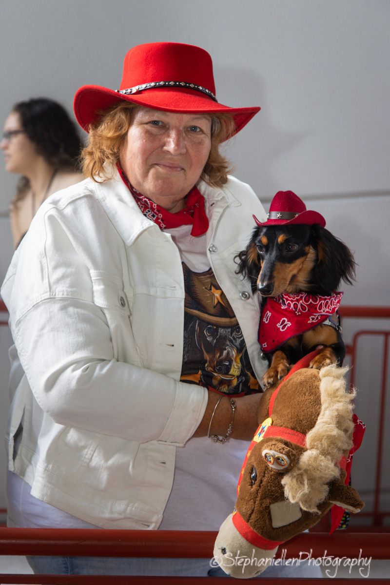 Woman in red cowboy hat holds dachshund dressed in matching cowboy costume with red bandana at dog event.