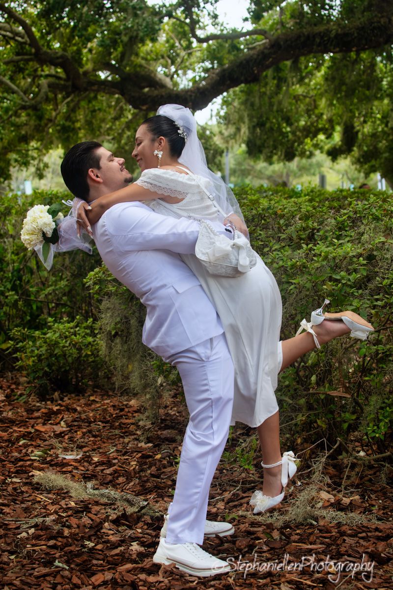 Groom lifting bride in white wedding dress among lush green hedges and trees outdoors.