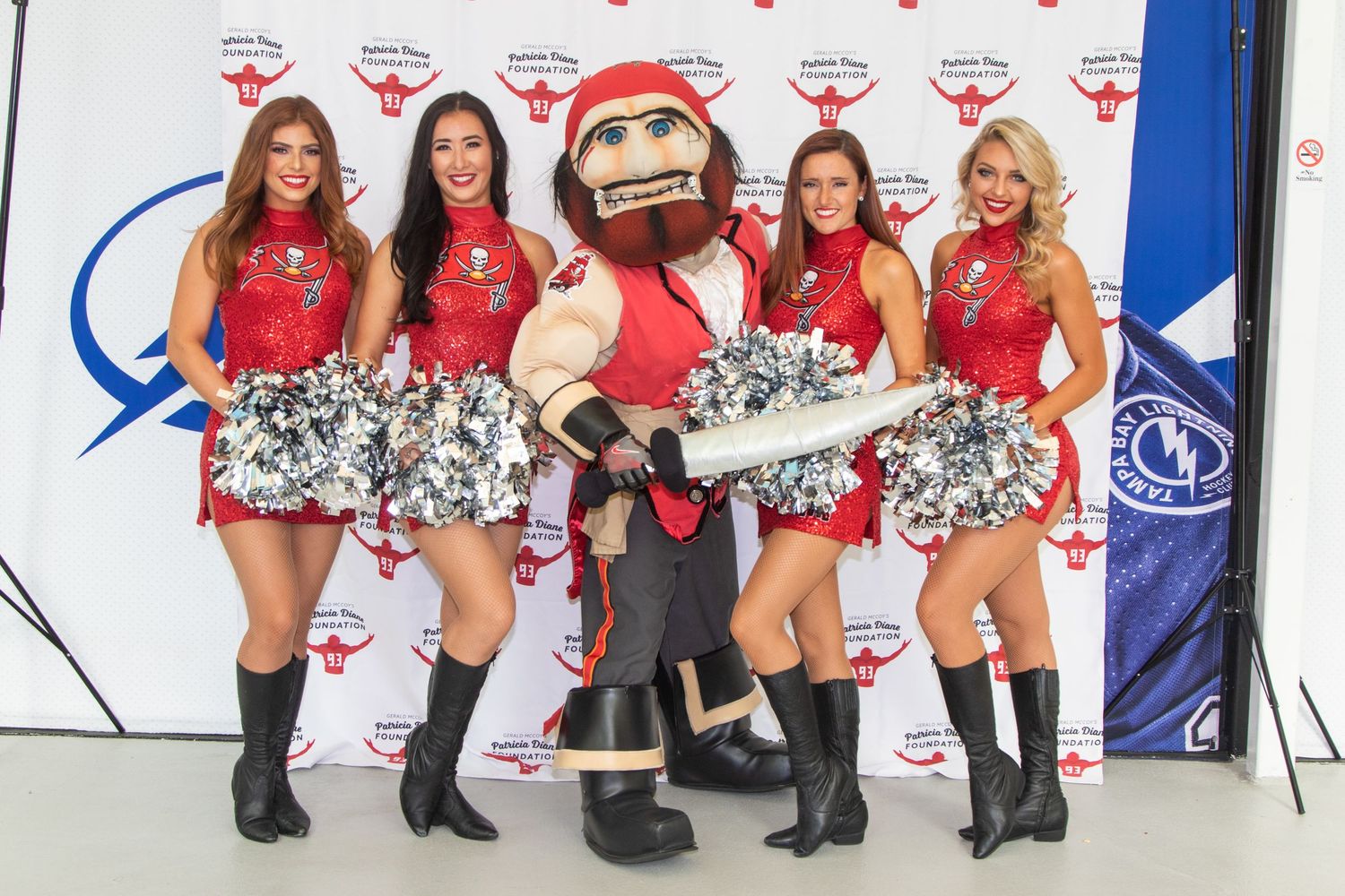 Four cheerleaders in red uniforms pose with a pirate mascot holding pom-poms at a sports event.