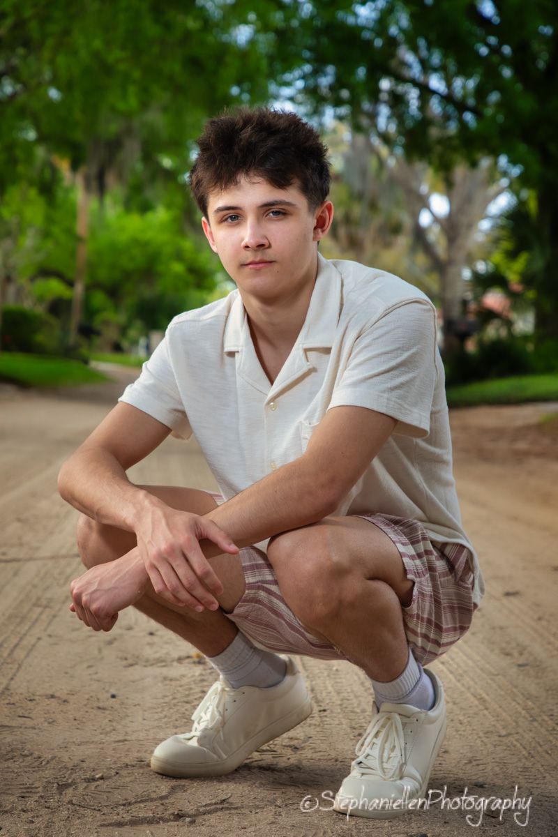 Young teenage boy crouching on a dirt path in a white polo shirt and plaid shorts outdoors.