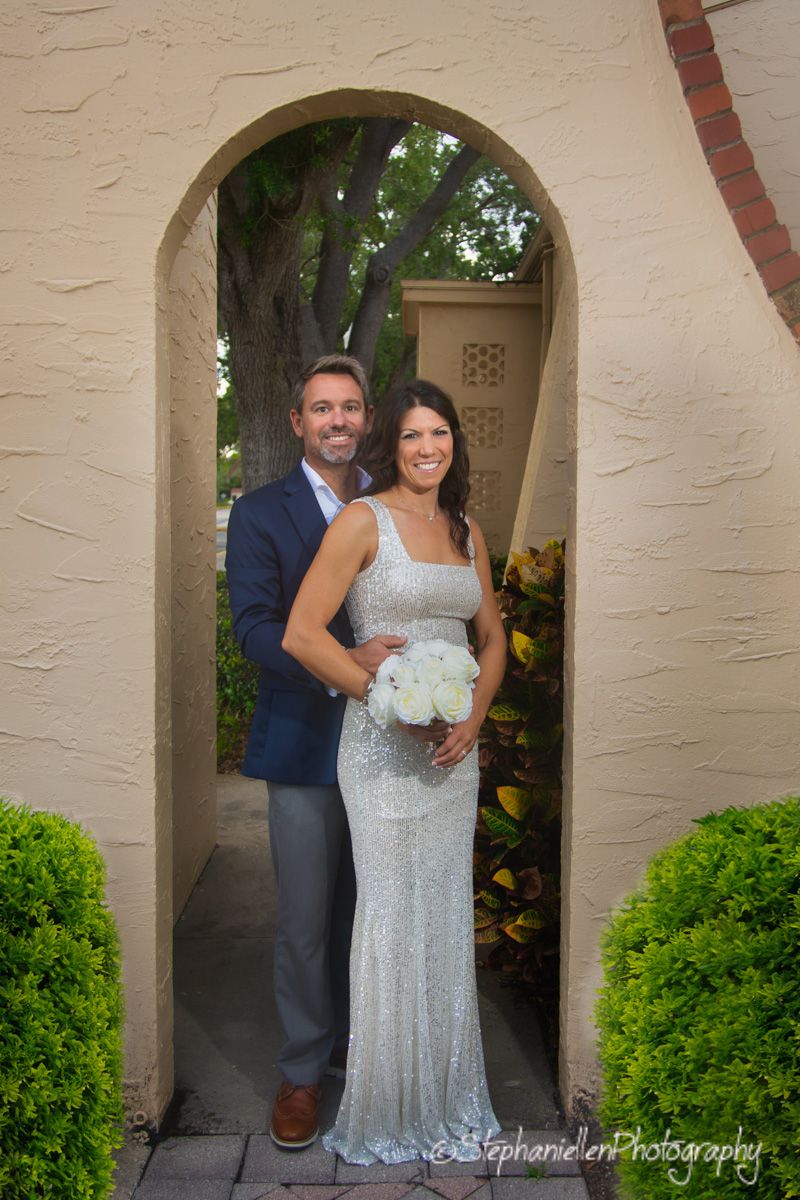 Smiling bride in silver gown holding white bouquet with groom in blue suit under stone arch.