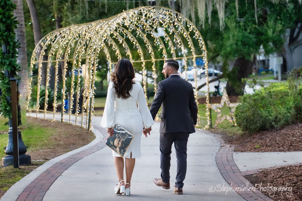 Couple holding hands walking down a pathway under a canopy of twinkling lights at a romantic outdoor wedding venue.