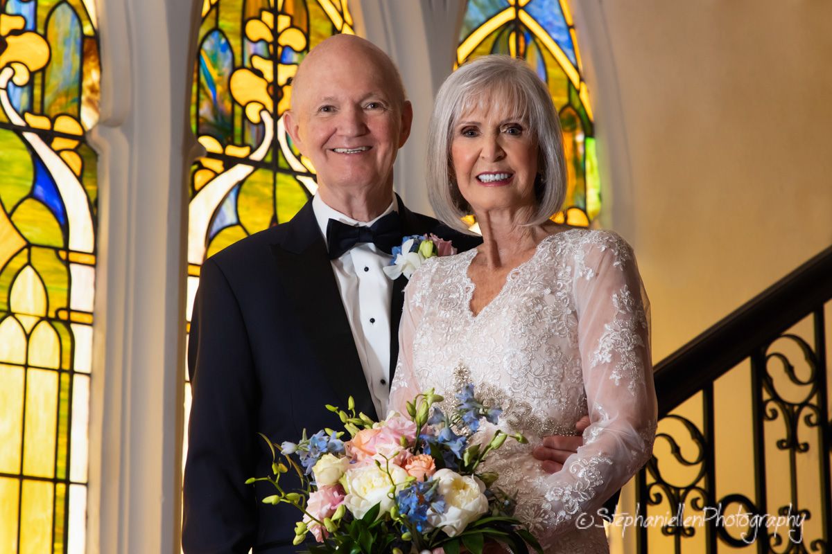 Elegant older bride and groom pose on church stairs before colorful stained glass window.