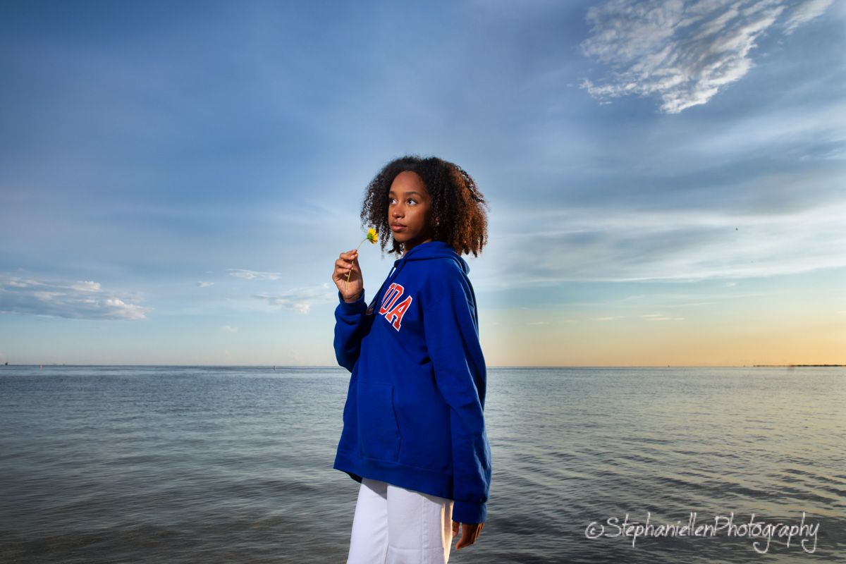 Young woman in royal blue jacket posing dramatically by calm ocean water at sunset.
